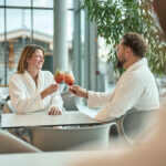 Paar sitzt in Bademänteln in der Thermenlounge und stößt mit Coktails an.