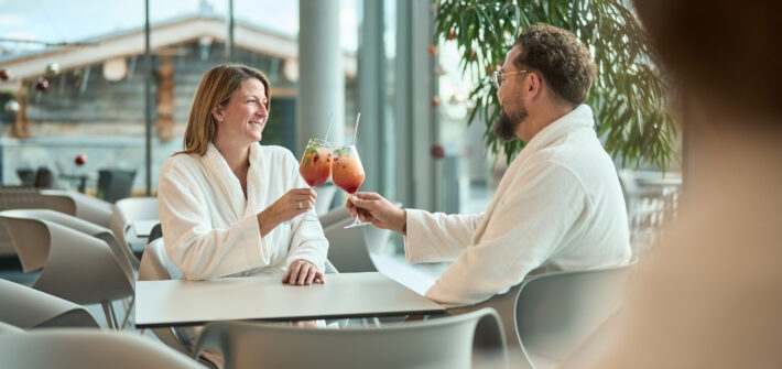 Paar sitzt in Bademänteln in der Thermenlounge und stößt mit Coktails an.