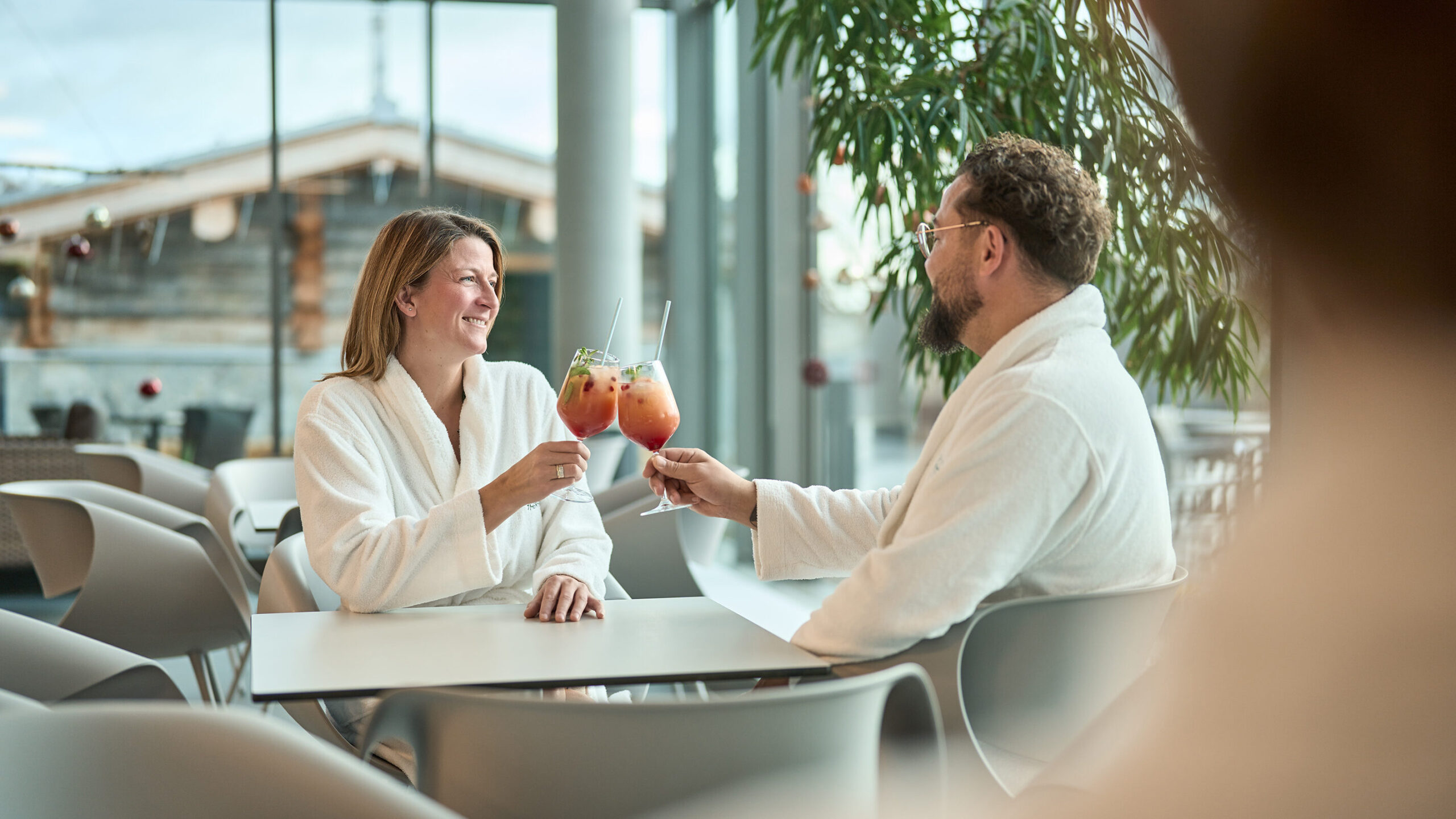 Paar sitzt in Bademänteln in der Thermenlounge und stößt mit Coktails an.