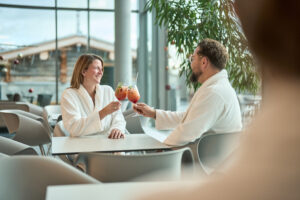 Paar sitzt in Bademänteln in der Thermenlounge und stößt mit Coktails an.